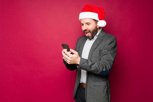Emocionado hombre con un sombrero de santa recibir un mensaje de texto sorprendente o el uso de las redes sociales durante las vacaciones de Navidad - Foto, imagen
