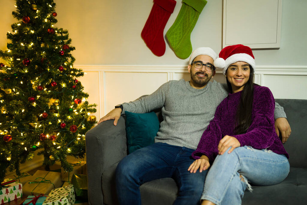 Ελκυστικό νεαρό ζευγάρι με καπέλα Σάντα αγκαλιάζει ενώ κάθεται στον καναπέ δίπλα στο χριστουγεννιάτικο δέντρο - Φωτογραφία, εικόνα