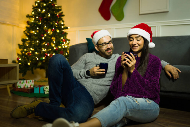 Pareja cariñosa con sombreros de santa disfrutando de una noche de Navidad en casa bebiendo chocolate caliente al lado del árbol - Foto, Imagen