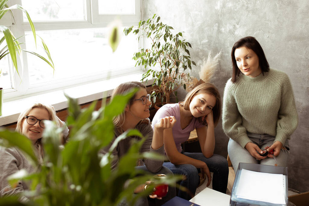 Positive, smiling, tired women sit together, packing luggage in living room. Relocation, moving in to new place. Sitting and waiting for shipmen, pack paper folders and house plants. New chapter - 写真・画像