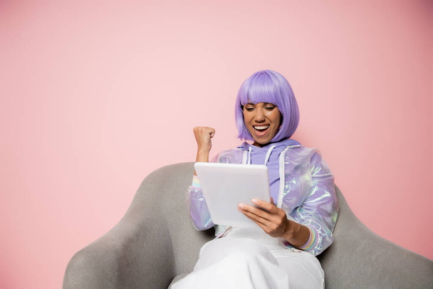 excited african american woman in purple wig using digital tablet and sitting in armchair isolated on pink  - Fotó, kép