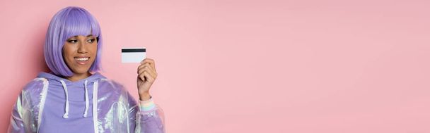 šťastná africká americká žena s fialovými vlasy držící kreditní kartu izolované na růžové, banner - Fotografie, Obrázek