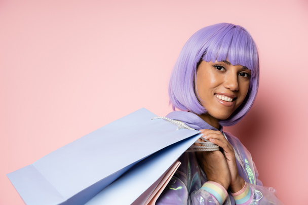 joyful and young african american woman with purple hair holding shopping bags on pink  - Fotografie, Obrázek