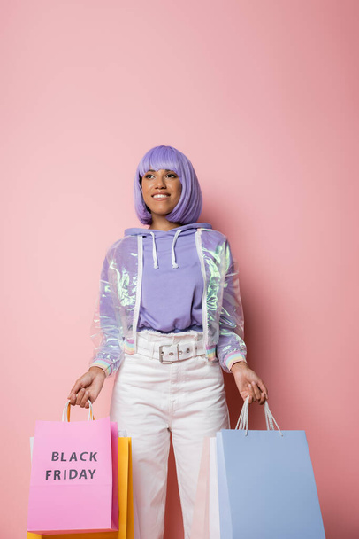 joyful african american woman with purple hair holding shopping bags with black friday lettering on pink  - Zdjęcie, obraz