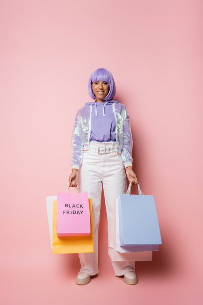 alegre mujer afroamericana con pelo morado sosteniendo bolsas de compras con letras de viernes negro en rosa  - Foto, Imagen