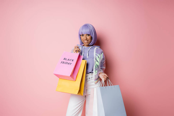 positive african american woman with purple hair holding shopping bags with black friday lettering on pink  - Photo, Image