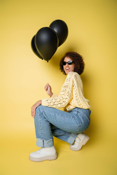 full length of cheerful african american woman in sunglasses holding black balloons while sitting on yellow - Foto, Imagen