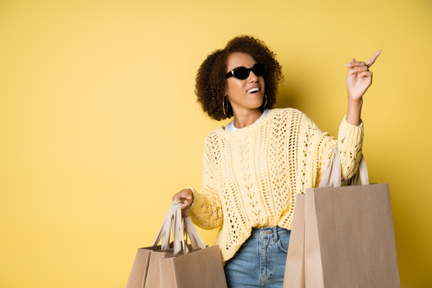 šťastná africká Američanka ve stylových slunečních brýlích držící nákupní tašky a ukazující prstem na žlutou - Fotografie, Obrázek