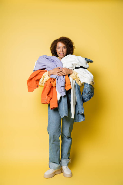full length of happy african american woman holding pile of colorful clothes on yellow  - Foto, Imagen