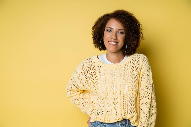 happy and young african american woman in knitted sweater posing with hand on hip on yellow - Zdjęcie, obraz