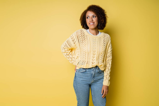 cheerful and young african american woman in knitted sweater posing with hand on hip on yellow - Photo, Image