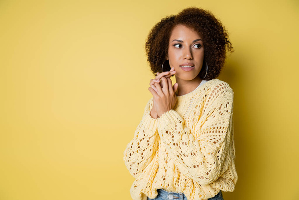 curieuse et jeune femme afro-américaine en pull tricoté regardant loin sur jaune - Photo, image