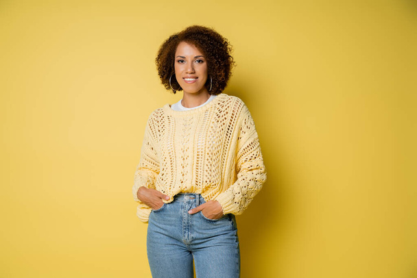 fröhliche junge afrikanisch-amerikanische Frau im Strickpullover stehend mit den Händen in den Taschen auf Jeans auf gelb - Foto, Bild