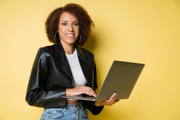 mulher americana africana feliz e jovem em jaqueta de couro usando laptop em amarelo - Foto, Imagem