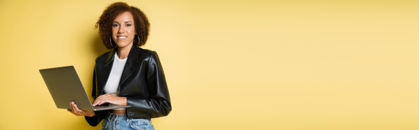 黄色のバナーのラップトップを使用して革のジャケットで幸せと若いアフリカ系アメリカ人女性 - 写真・画像