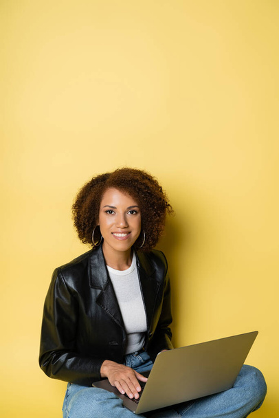 joyful and young african american woman in leather jacket using laptop on yellow - Фото, изображение