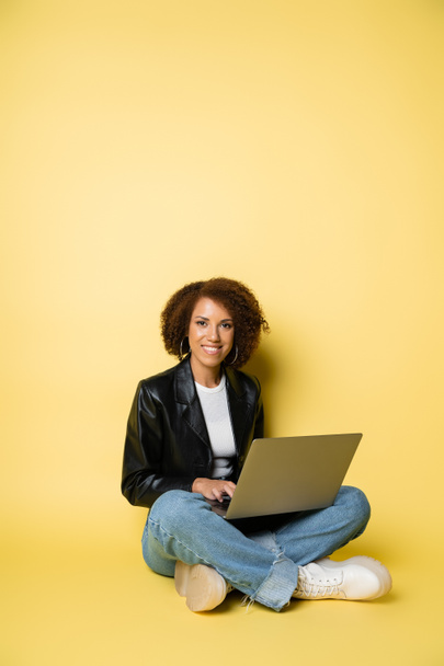 radostné a mladé africké americké ženy v kožené bundě pomocí notebooku, zatímco sedí na žluté - Fotografie, Obrázek