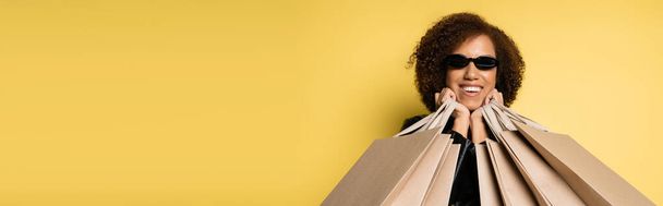 femme afro-américaine gaie dans des lunettes de soleil tenant des sacs à provisions le vendredi noir sur jaune, bannière - Photo, image