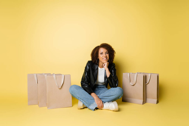 Deri ceketli ve kot pantolonlu mutlu Afro-Amerikan kadın alışveriş torbalarının yanında oturuyor. - Fotoğraf, Görsel