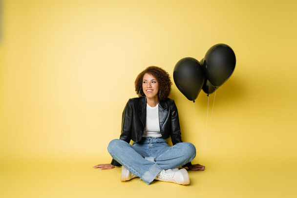 full length of smiling african american woman in leather jacket and jeans sitting near black balloons on yellow - Фото, зображення