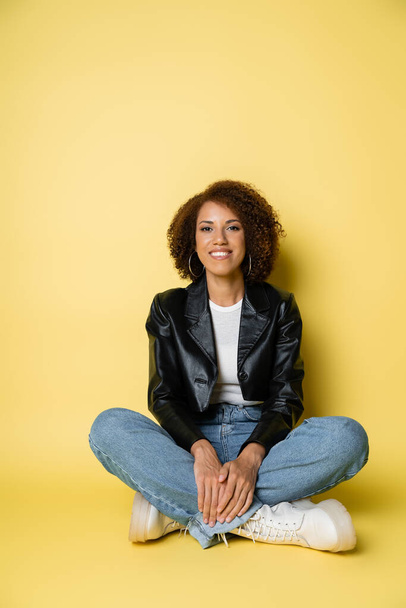 full length of pleased african american woman in leather jacket and jeans sitting with crossed legs on yellow - Φωτογραφία, εικόνα