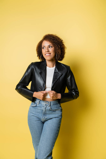 pleased african american woman in stylish leather jacket and jeans posing on yellow - Fotografie, Obrázek