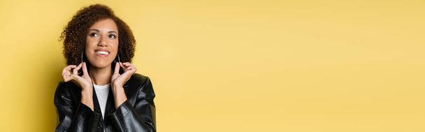 happy african american woman in stylish leather jacket touching hoop earrings on yellow, banner - Foto, Imagem