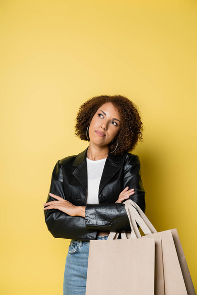 pensive african american woman in stylish leather jacket holding shopping bags with purchases on yellow - Foto, Bild