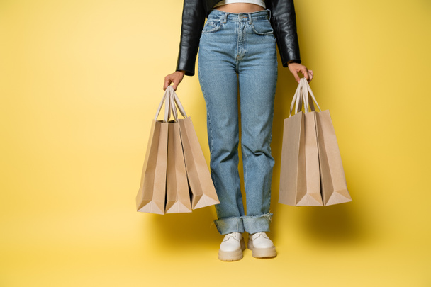 cropped view of african american woman in leather jacket and jeans holding shopping bags with purchases on yellow - Φωτογραφία, εικόνα