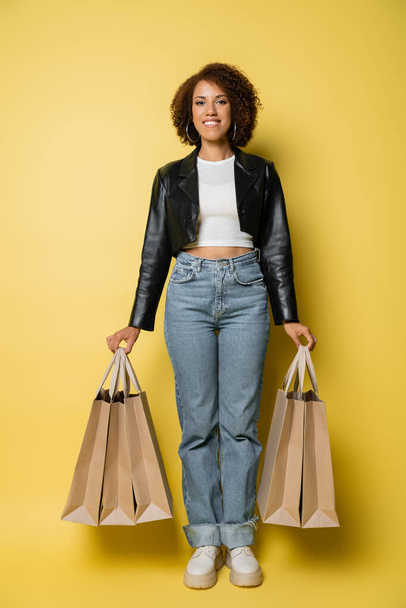 plná délka usmívající se africké Američanky ve stylové kožené bundě držící nákupní tašky s nákupy na žluté - Fotografie, Obrázek