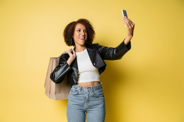mujer afroamericana feliz en elegante chaqueta de cuero sosteniendo bolsas de compras y tomando selfie en amarillo - Foto, imagen