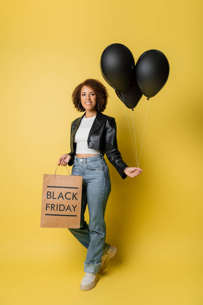 full length of smiling african american woman in leather jacket holding black friday shopping bag and balloons on yellow - Zdjęcie, obraz