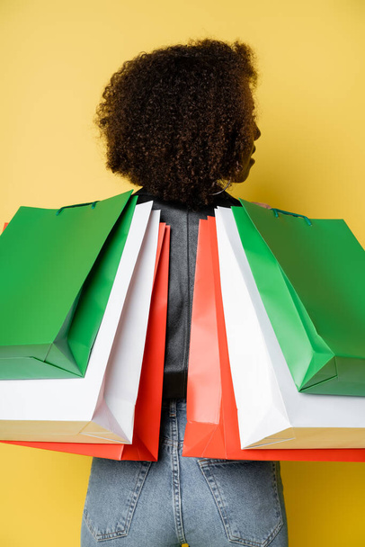 curly african american woman in denim jeans holding colorful black friday shopping bags on yellow - Photo, Image