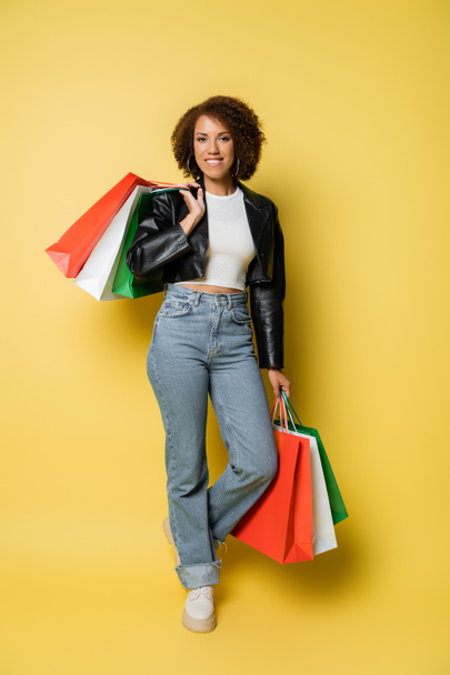 full length of positive african american woman in jeans and leather jacket holding colorful black friday shopping bags on yellow  - Fotó, kép
