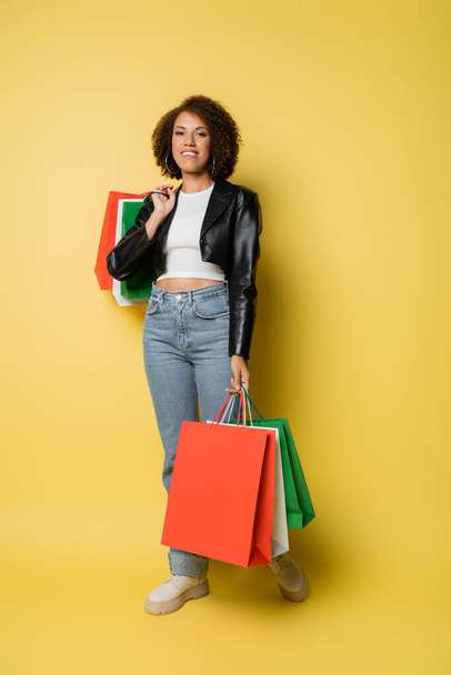 full length of cheerful african american woman in jeans and leather jacket holding colorful black friday shopping bags on yellow  - Zdjęcie, obraz