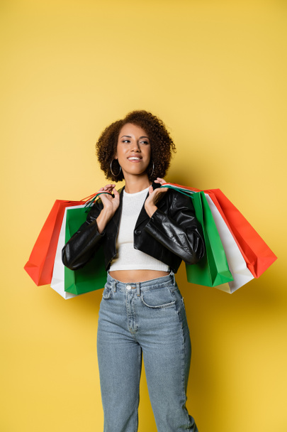 smiling african american woman in jeans and leather jacket holding colorful black friday shopping bags on yellow  - Photo, Image