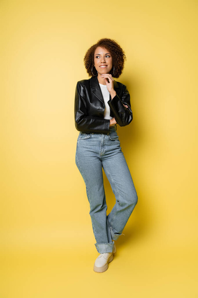 full length of smiling african american woman in jeans and leather jacket posing on yellow  - Fotografie, Obrázek