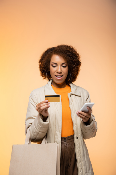 glimlachende Afrikaans-Amerikaanse vrouw in jas met smartphone en creditcard, terwijl staande met boodschappentas op oranje  - Foto, afbeelding