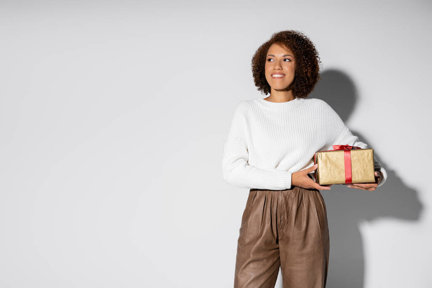 happy african american woman in autumnal outfit holding wrapped gift box on grey - Photo, Image