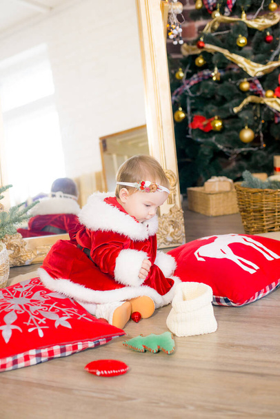 A little girl under one year old dressed as Santa Claus in a room decorated for Christmas, near the Christmas tree among the pillows, gifts, garlands and pine needles. Christmas mood. Children and Christmas - Фото, изображение