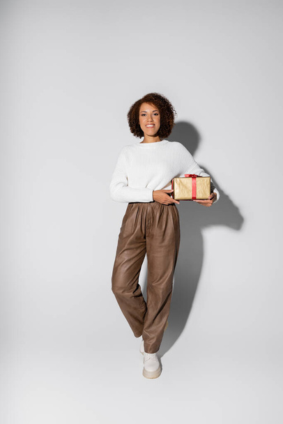 full length of smiling african american woman in autumnal outfit holding wrapped gift box on grey - Foto, Bild