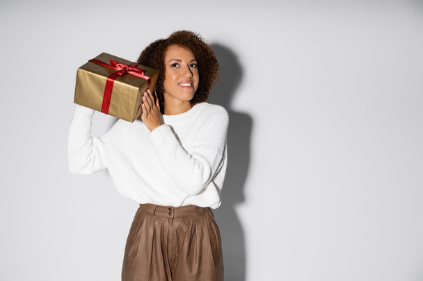 heureuse femme afro-américaine en tenue automnale tenant boîte cadeau enveloppée près de l'oreille sur gris - Photo, image