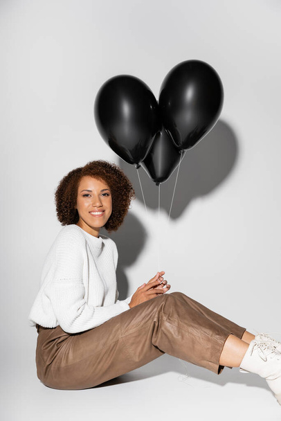 cheerful african american woman in autumnal outfit holding black balloons on grey  - Photo, image