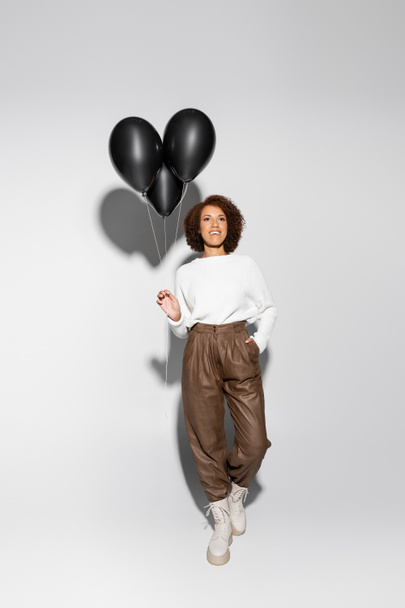 full length of smiling african american woman in autumnal outfit holding black balloons and posing with hand in pocket on grey - Zdjęcie, obraz