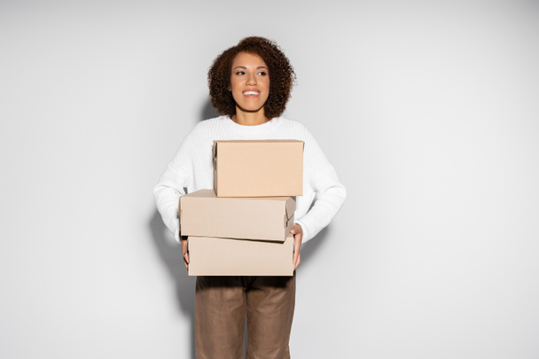 fröhliche afrikanisch-amerikanische Frau mit lockigem Haar hält Lieferboxen auf grau - Foto, Bild