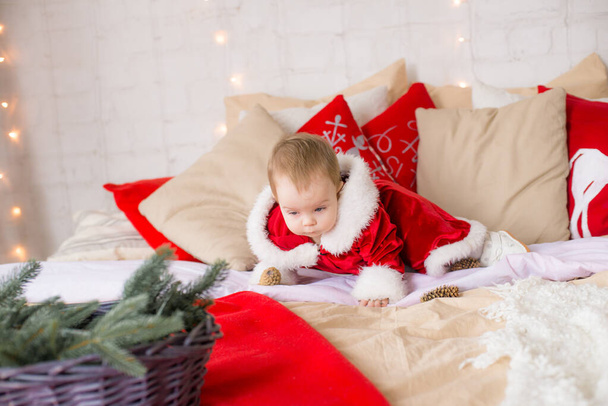 A little girl under one year old dressed as Santa Claus on a large bed in a room decorated for Christmas, among the pillows of garlands and pine needles. Christmas mood. Children and Christmas - Фото, изображение