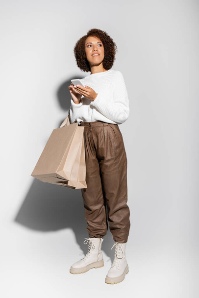 full length of happy african american woman in autumnal outfit holding shopping bags and using smartphone on grey - Foto, Bild