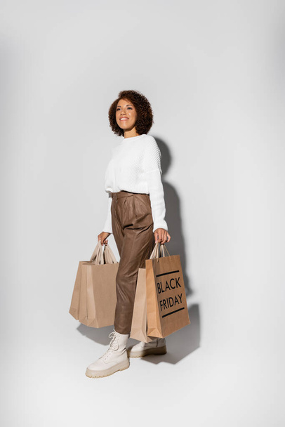 pleased african american woman in autumnal outfit holding shopping bags with black friday lettering on grey - Photo, Image