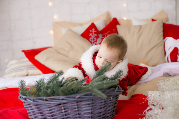A little girl under one year old dressed as Santa Claus on a large bed in a room decorated for Christmas, among the pillows of garlands and pine needles. Christmas mood. Children and Christmas - 写真・画像
