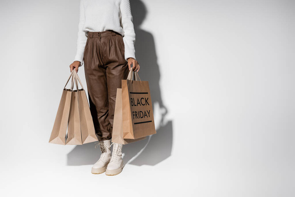 partial view of african american woman in autumnal outfit holding shopping bags with black friday lettering on grey - Fotoğraf, Görsel